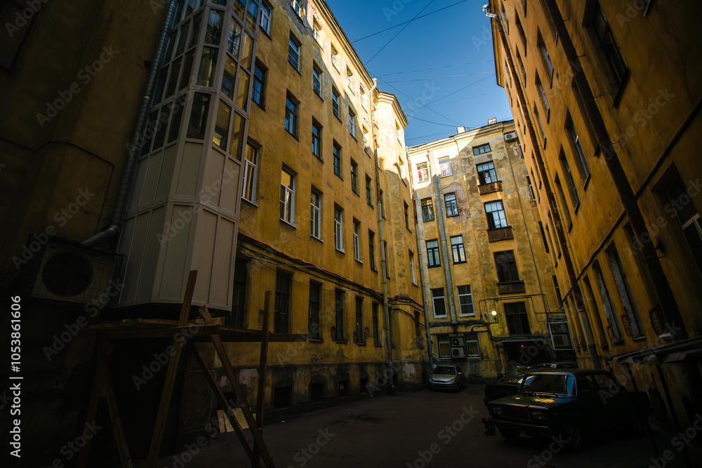 Fototapeta premium A view from the old Saint Petersburg courtyard, looking up towards the sky, framed by worn-out, weathered walls and parked cars below.