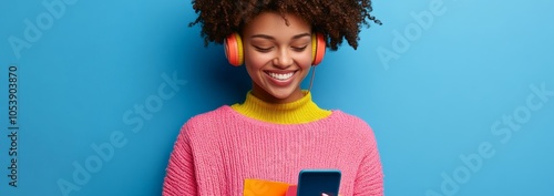 Beautiful image of a stylish young woman wearing knitted sweater, pink hat on blue background, listening to music in headphones and holding her smartphone while holding shopping bags