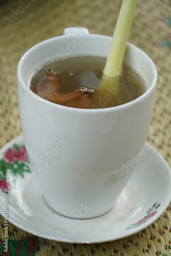 A cup of hot ginger tea (Zingiber officinale) with sugar and lemongrass (Cymbopogon citratus), served in a white cup and saucer. A popular Indonesian drink. Blurred woven mat background.