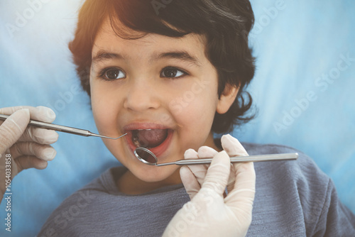 Cute arab boy sitting at dental chair with open mouth during oral checking up with doctor. Visiting sunny dentist office
