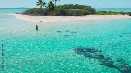 Aerial view of a young woman walking on the shallow water of a tropical island with turquoise water and a small island with palm trees in the background