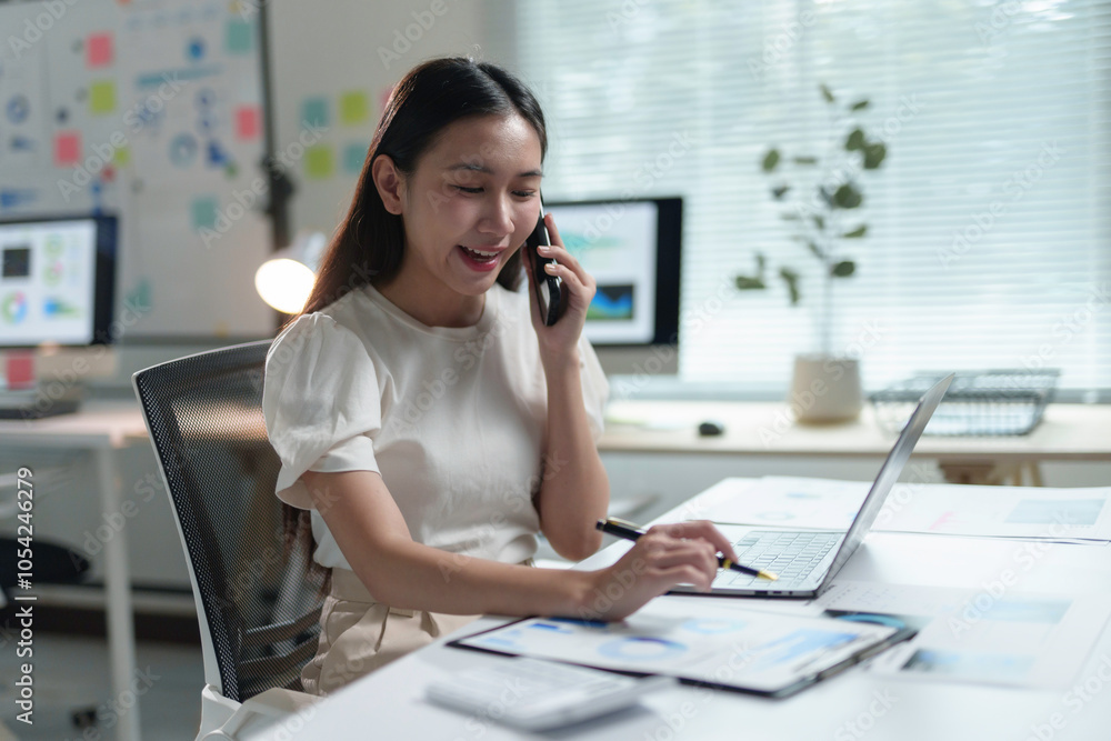 Fototapeta premium Smiling businesswoman working with documents and laptop while talking on the phone in modern office