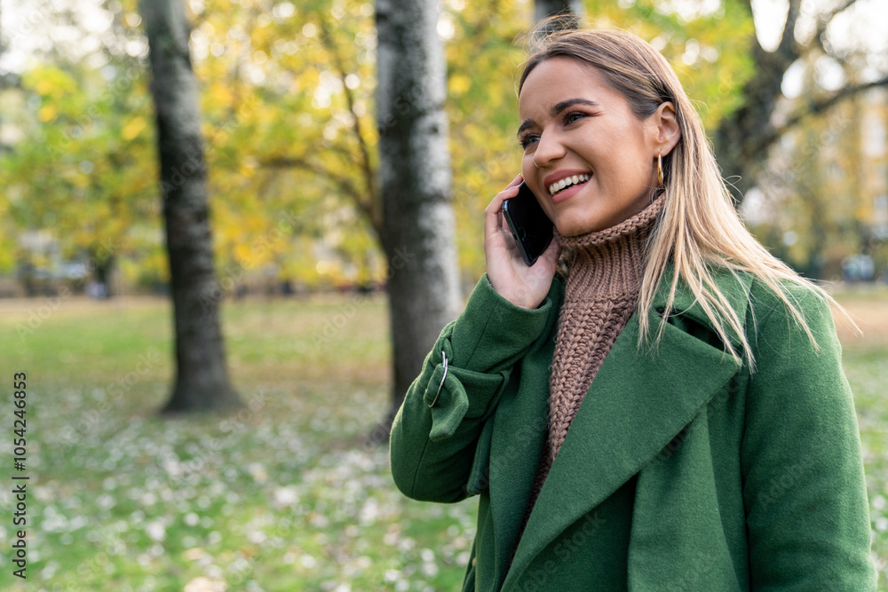 A joyful Caucasian woman engages with her smartphone during a leisurely stroll, adorned in a cozy sweater and chic green coat, surrounded by fall foliage. A joyful Caucasian woman engages with her smartphone during a leisurely stroll, adorned in a cozy sweater and chic green coat, surrounded by fall foliage.