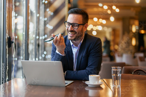 Mid adult Caucasian man in smart attire multitasks with a laptop and smartphone, comfortably seated in a well-lit modern cafe. photo