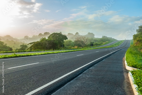 Paved dual-lane highway in perspective, with no cars, early in the morning, with a bit of fog and lush green landscape in the background in Brazil.