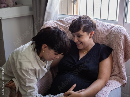 In a cozy room, an expectant mother smiles warmly while her wife gently touches her growing belly. The atmosphere is filled with love and anticipation for their new arrival. photo