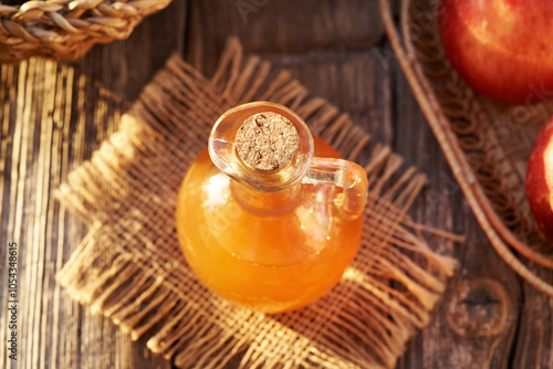 A jug of apple cider vinegar with red apples on a table in sunlight