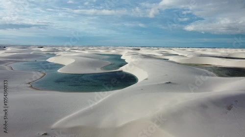 A drone reveals the vast white dunes of Lençóis Maranhenses, with shimmering lagoons nestled between the rolling sands. The sweeping aerial view captures the stunning contrast of the endless desert.