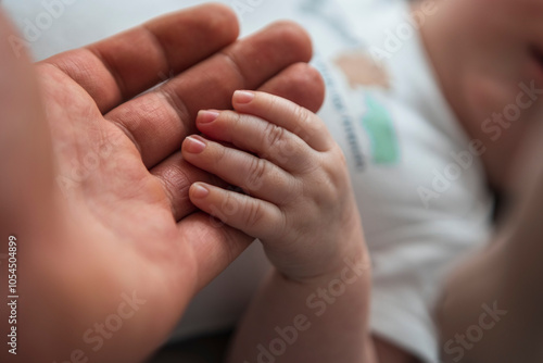 A heartwarming close-up of an adult’s hand gently holding a baby’s hand, capturing the tender connection between them. The contrast between the soft, delicate baby fingers and the larger photo