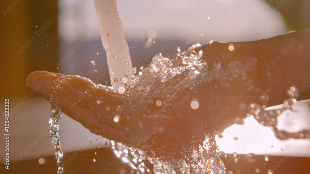 Fototapeta premium CLOSE UP, LENS FLARE: Crystal clear water splashes over an open hand beneath a stainless steel kitchen faucet, with morning sunlight illuminating each droplet. Fresh running water in modern household.