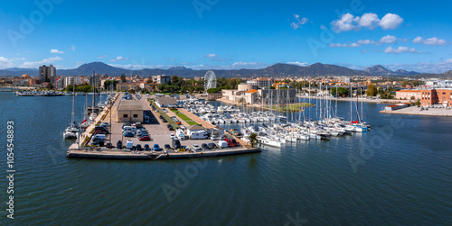 The vibrant marina in Olbia, Sardinia, features yachts and boats, a Ferris wheel, and a mix of modern and traditional architecture against rolling hills. photo