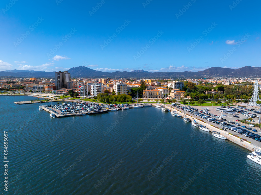 Fototapeta premium Aerial view of Olbia, Sardinia, featuring a marina with boats, a parking area, and a skyline of modern and traditional buildings against hills and mountains.