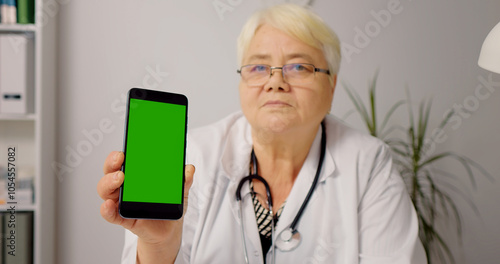 Selecive focus on phone with green screen mock up hold by mature doctor in her office. Woman doctor with stethoscope around her neck showing a phone with chroma key screen. photo