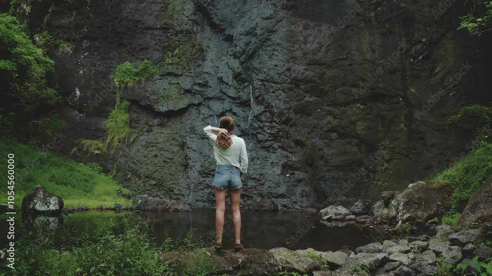 Tourist admiring a waterfall flowing over a rocky cliff in a tropical forest. She is enjoying a moment of peace and tranquility surrounded by lush vegetation Tourist admiring a waterfall flowing over a rocky cliff in a tropical forest. She is enjoying a moment of peace and tranquility surrounded by lush vegetation
