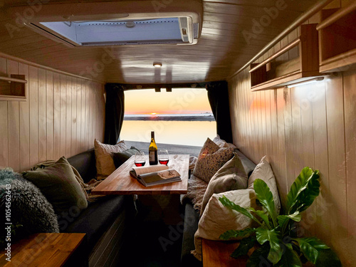 A warm and inviting camper van interior with plush seating, a wooden table, and wine glasses, set against a mesmerizing ocean sunset. Perfect for travel and leisure enthusiasts. photo