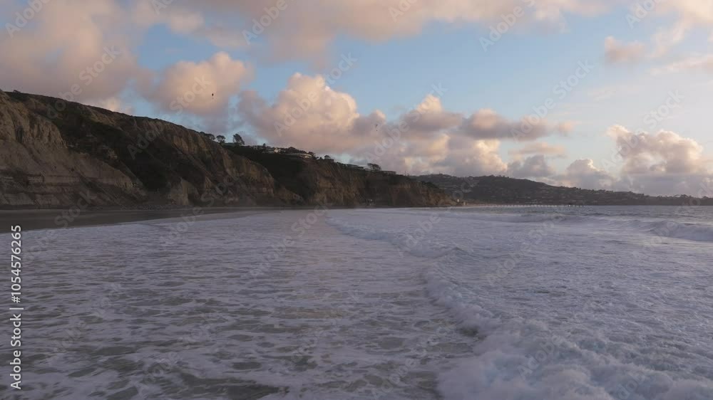Aerial view flying along the coast of Blacks Beach in La Jolla California with views of the Pacific Ocean waves crashing along the sandy shores and the rocky cliffs during golden hour sunset Aerial view flying along the coast of Blacks Beach in La Jolla California with views of the Pacific Ocean waves crashing along the sandy shores and the rocky cliffs during golden hour sunset