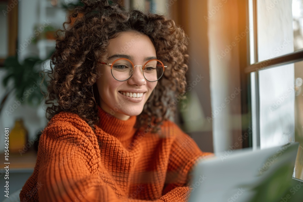 Happy woman with curly hair wearing glasses and orange sweater using laptop at desk watching online video in soft daylight home interior Happy woman with curly hair wearing glasses and orange sweater using laptop at desk watching online video in soft daylight home interior