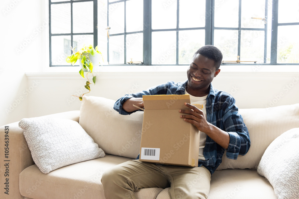 Φωτογραφία Smiling African american man opening package on couch at home, enjoying holiday