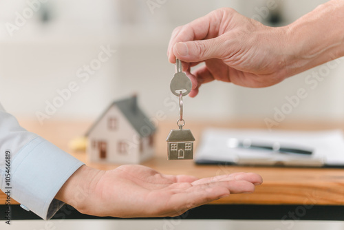 A hand passes house keys to another, symbolizing the transfer of property ownership, with a miniature house in the background.