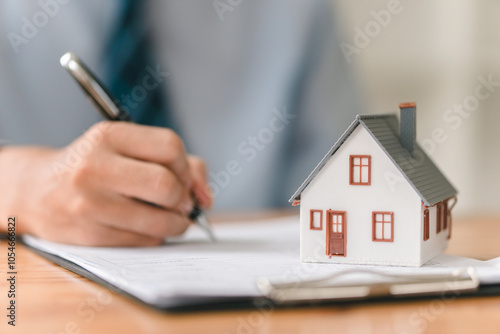 A person writes on a document next to a miniature house, symbolizing real estate transactions or home ownership activities.