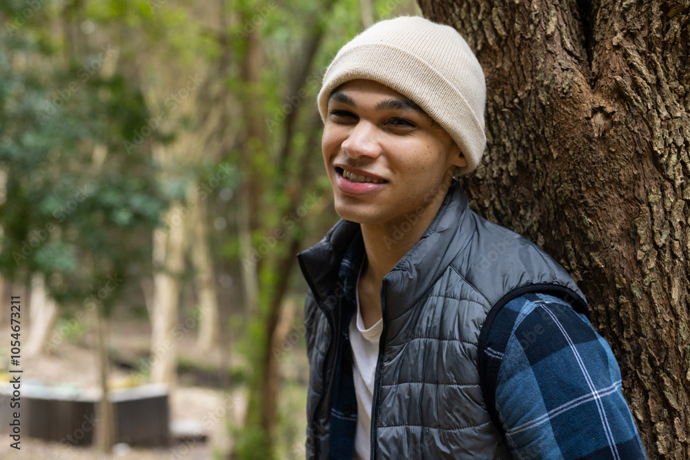 Smiling young man in beanie leaning against tree in forest, copy space Smiling young man in beanie leaning against tree in forest, copy space