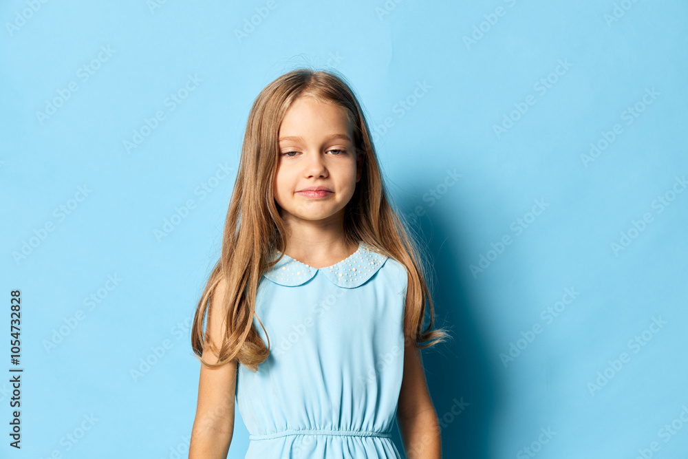 Confident little girl in a blue dress standing with hands on hips against matching blue background, expressing determination Confident little girl in a blue dress standing with hands on hips against matching blue background, expressing determination