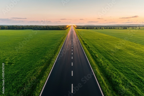 Long stretch of empty road with green fields under a colorful sunset sky.