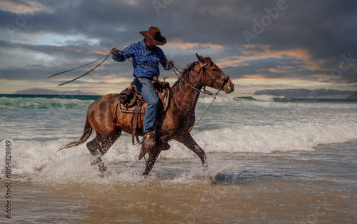A cowboy riding a horse in the water causing splashes at a beach with a dramatic sky The image is from Rosarito Mexico