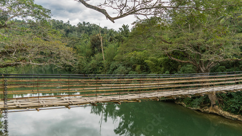 A rickety Bamboo Hanging Bridge spans over a tropical river. You can see the weaving of the footpath, the railing. Lush green vegetation on the shores. Reflection on calm water. Philippines.  Bohol  photo