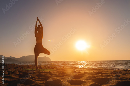 Vrikshasana tree pose from yoga, female silhouette at sunset or sunrise. Back view of a girl practicing morning workout on the seashore photo