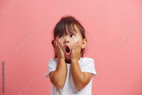 Amazed little asian girl in white t shirt shocked keeps hand on cheek, looks up, expresses surprisement, isolated over pink background.  photo