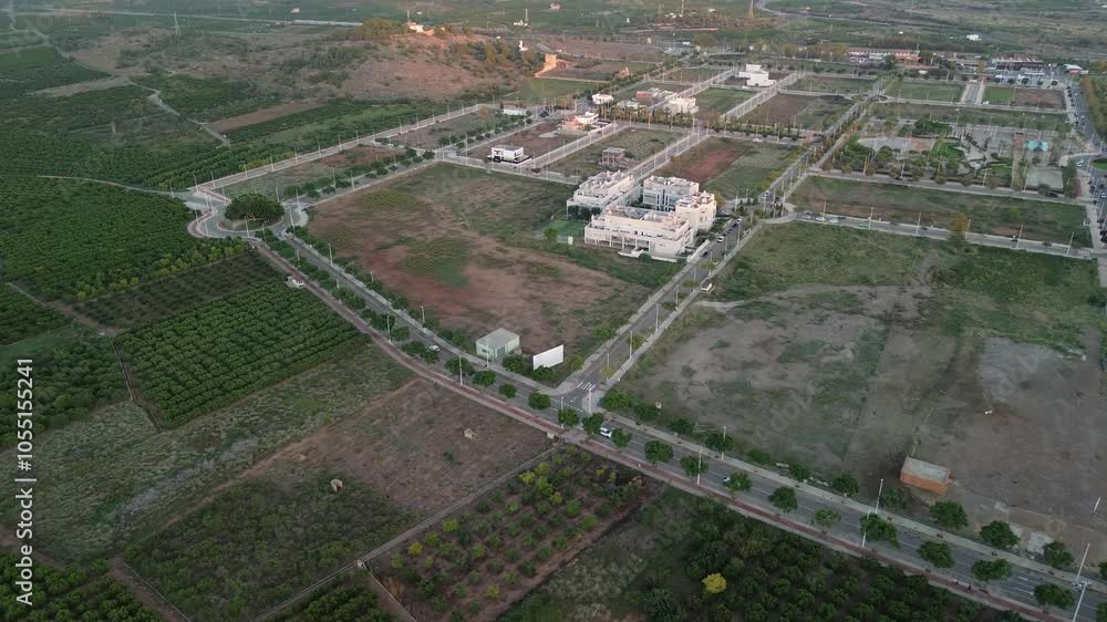 Aerial shot capturing a suburban development surrounded by lush green fields. Shows a planned layout of roads, residential areas, and undeveloped land