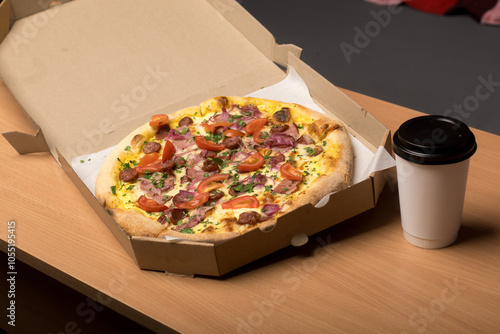 Open pizza box with a freshly baked pizza topped with tomatoes, sausage, ham, and herbs on a wooden table. A takeaway coffee cup is placed next to it. Perfect for food delivery and casual dining theme