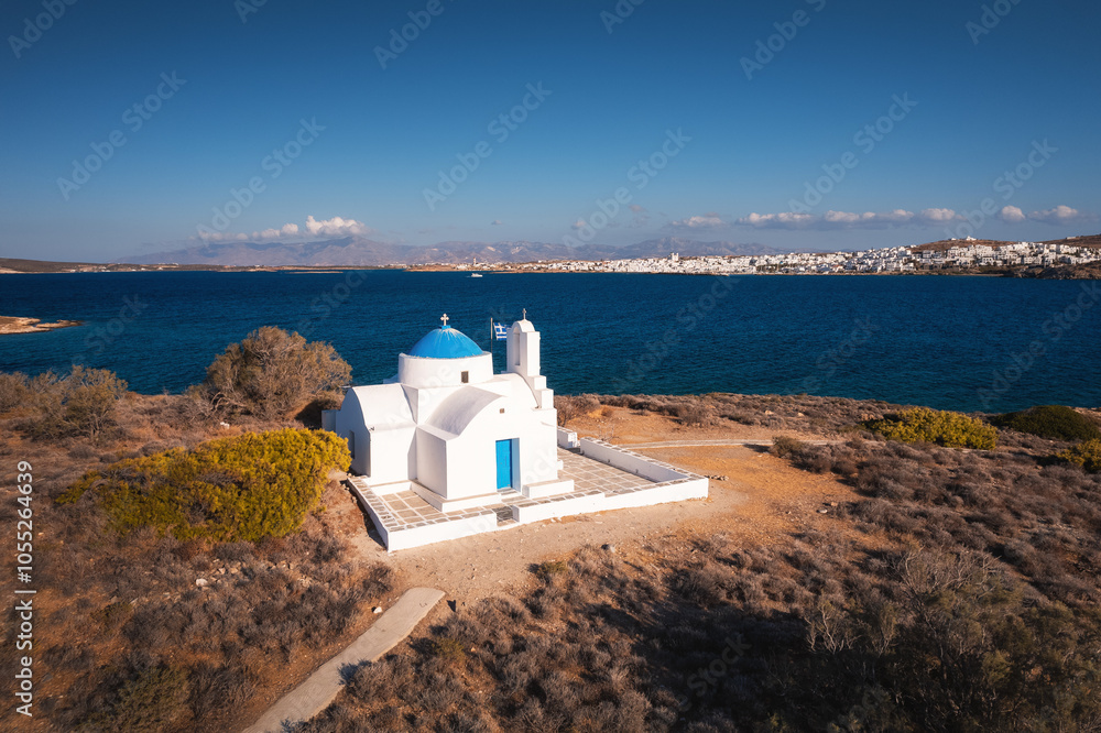 A charming blue-domed chapel on Paros Island, Greece, set against the backdrop of the shimmering Aegean Sea. The whitewashed walls, vibrant dome, and surrounding stone path create a picturesque scene