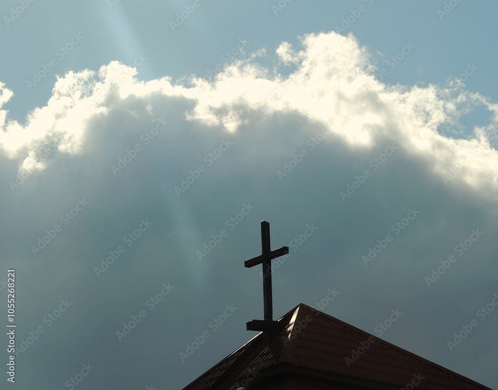 simple dark cross on top of protestant church roof against stormy beautiful sky, concept of faith in god against contrast of dark cloud against shining sky with cross in center