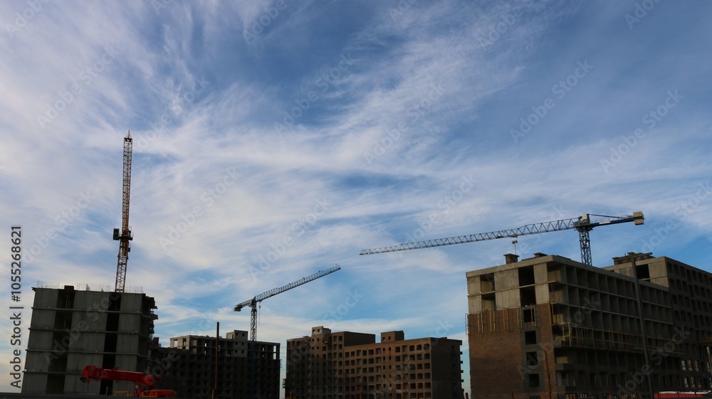 Three construction cranes sit behind unfinished multi-story buildings against a blue cloudy sky Three construction cranes sit behind unfinished multi-story buildings against a blue cloudy sky