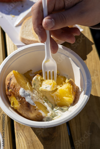 A close-up of a baked potato served in a disposable bowl with creamy herb sauce, being eaten with a plastic fork. A simple and comforting outdoor meal enjoyed on a wooden table.