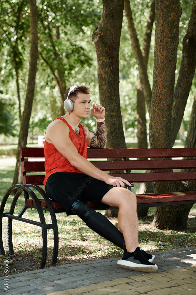 Fototapeta premium A young man with tattoos sits on a bench outdoors, enjoying the music in headphones.