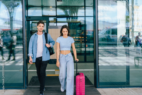 Two young adults exiting airport building, male and female, holding luggage. Man in casual outfit, woman in blue top, pink suitcase. Sunny day, travel setting, reflecting glass doors.