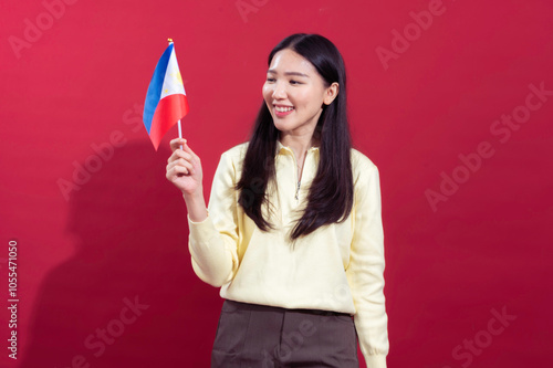 Asian woman smiling, wearing a yellow sweater, holding a small Philippine flag in her hand. She stands against a solid red background, exuding a sense of pride and joy