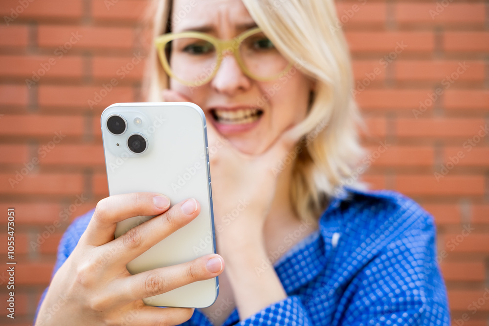 Fototapeta premium A woman with blond hair and bright glasses is looking at her smartphone with a shocked expression. She appears surprised or wonder while standing outdoor. 