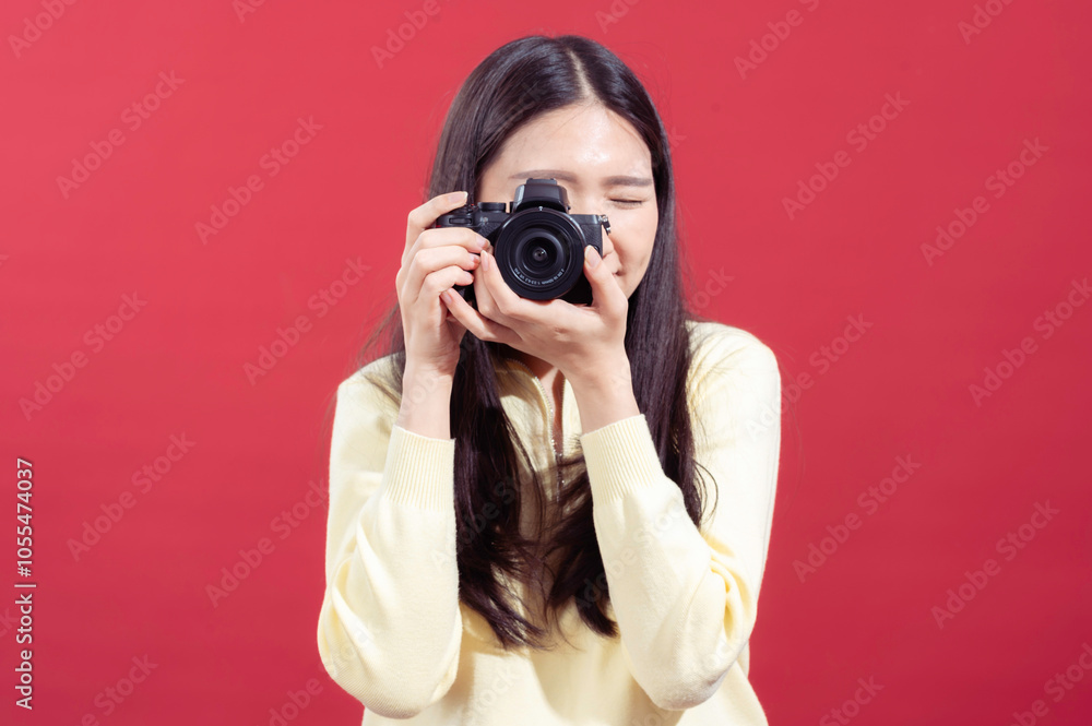 Asian woman in a yellow sweater holding a camera up to her face, as if taking a photo, with one eye closed. She is against a solid red background, creating a lively and focused scene Asian woman in a yellow sweater holding a camera up to her face, as if taking a photo, with one eye closed. She is against a solid red background, creating a lively and focused scene