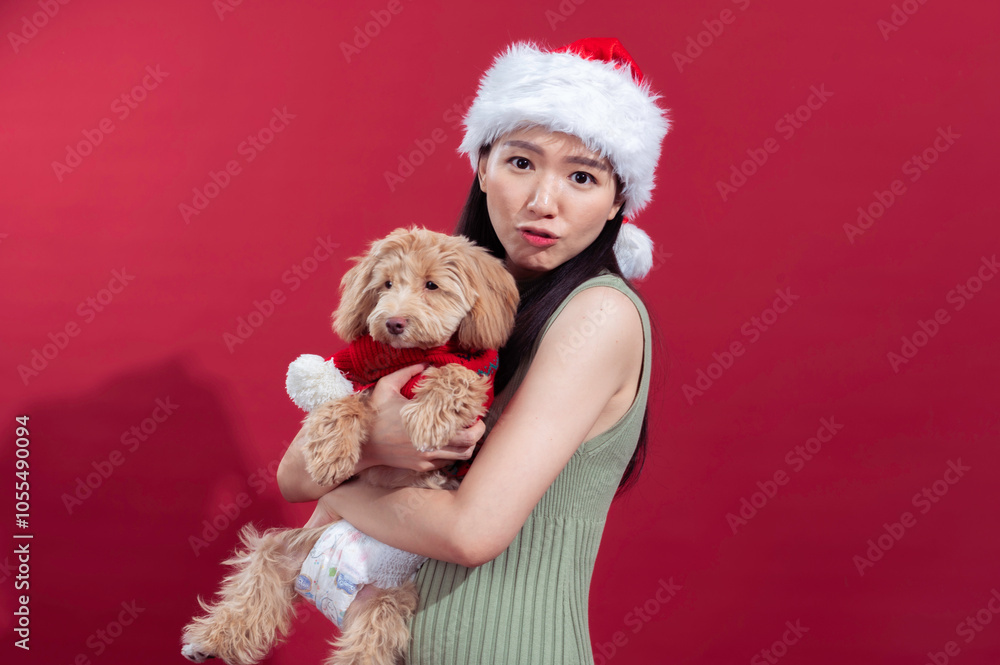 An Asian woman wearing a Santa hat and a sleeveless green dress is smiling while holding a small, fluffy dog dressed in a festive red outfit. They are standing in front of a bright red background An Asian woman wearing a Santa hat and a sleeveless green dress is smiling while holding a small, fluffy dog dressed in a festive red outfit. They are standing in front of a bright red background
