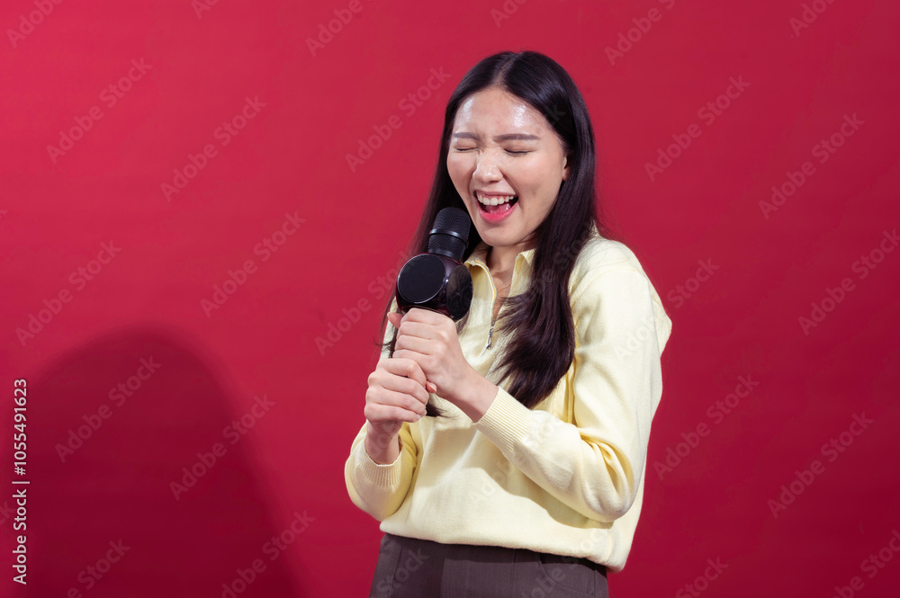 Asian woman smiling and singing into a microphone, wearing a yellow sweater with a red background. She appears enthusiastic and expressive, holding the microphone Asian woman smiling and singing into a microphone, wearing a yellow sweater with a red background. She appears enthusiastic and expressive, holding the microphone