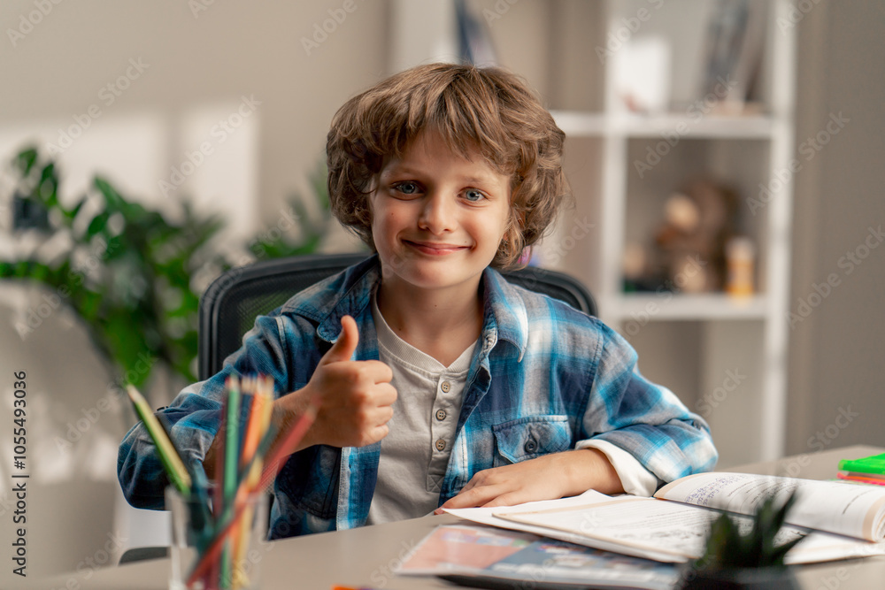 close up in the children's room a schoolboy in white sweater and a blue checkered shirt at a desk doing homework on his studies writing in a notebook looks at the camera close up in the children's room a schoolboy in white sweater and a blue checkered shirt at a desk doing homework on his studies writing in a notebook looks at the camera