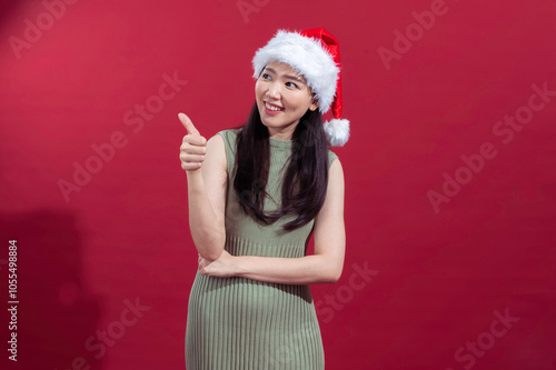 An Asian woman wearing a Santa hat and a sleeveless green dress gives a thumbs-up with a cheerful smile. She stands in front of a solid red background, conveying a festive, positive mood photo