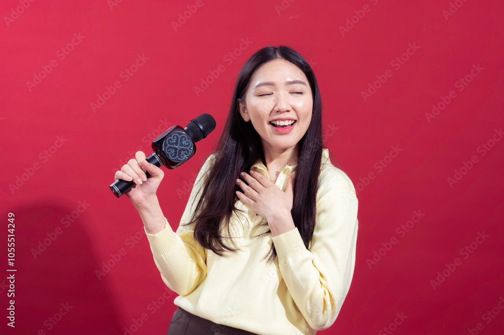 Asian woman smiling and singing into a microphone, wearing a yellow sweater with a red background. She appears enthusiastic and expressive, holding the microphone Asian woman smiling and singing into a microphone, wearing a yellow sweater with a red background. She appears enthusiastic and expressive, holding the microphone