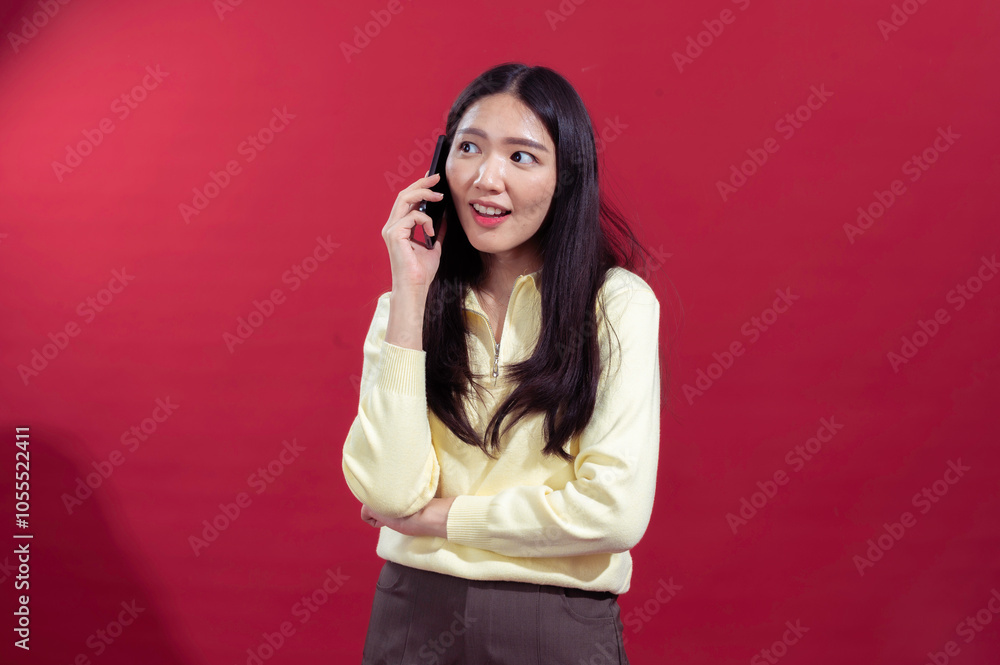 Asian woman smiling and holding a smartphone to her ear, dressed in a yellow sweater with a red background. The expression suggests she’s engaged in a cheerful phone conversation Asian woman smiling and holding a smartphone to her ear, dressed in a yellow sweater with a red background. The expression suggests she’s engaged in a cheerful phone conversation