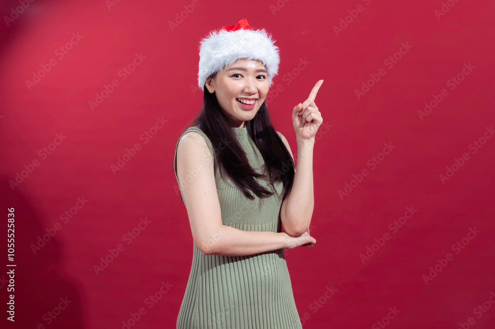 An Asian woman wearing a Santa hat, dressed in a sleeveless green dress, smiles brightly while raising her index finger, as if making a point, with a red background behind her An Asian woman wearing a Santa hat, dressed in a sleeveless green dress, smiles brightly while raising her index finger, as if making a point, with a red background behind her