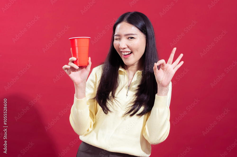 Asian woman smiling and holding a red plastic cup in her hand, wearing a yellow sweater against a red background. She looks cheerful and relaxed, with one arm crossed and the other holding cup Asian woman smiling and holding a red plastic cup in her hand, wearing a yellow sweater against a red background. She looks cheerful and relaxed, with one arm crossed and the other holding cup